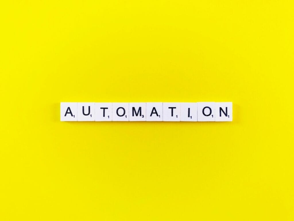 The image shows the word "AUTOMATION" spelled out with black letters on white square tiles, arranged in a horizontal line. The tiles are centered against a bright yellow background, creating a striking and high-contrast visual. © WEB6 Cloud