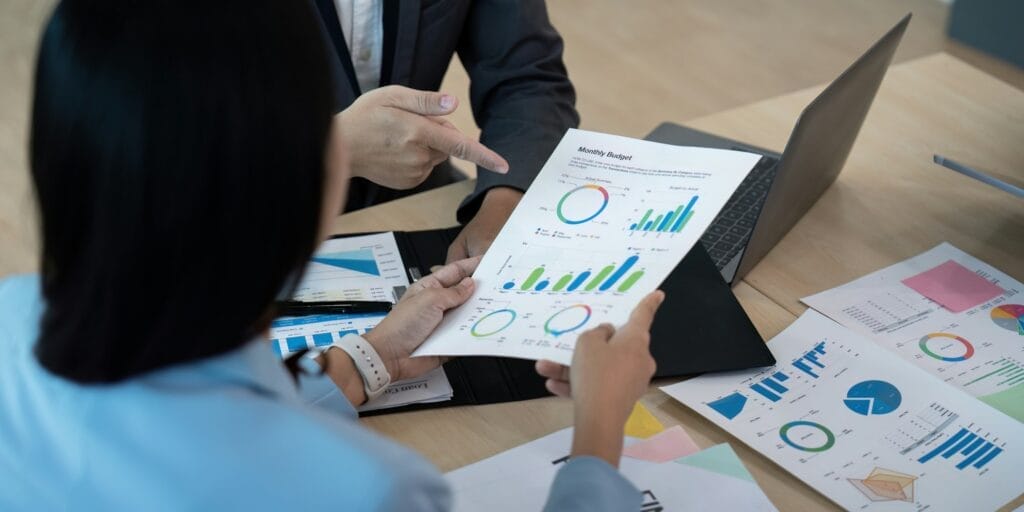 Two people are reviewing a printed Monthly Budget report filled with colorful charts and graphs at a desk, engaging in an AI-assisted decision-making process. One person is pointing to the report while the other holds it. A laptop, additional documents with charts, and colorful papers on the desk suggest a financial or business discussion. © WEB6 Cloud
