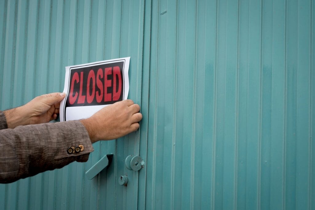 A person with a brown plaid jacket and buttoned sleeves is posting a sign that reads "CLOSED" in bold red letters over a white background. The sign is being attached to the middle of a teal corrugated metal door, suggesting the company is closing due to obsoleteness amid the Artificial Intelligence revolution. © WEB6 Cloud
