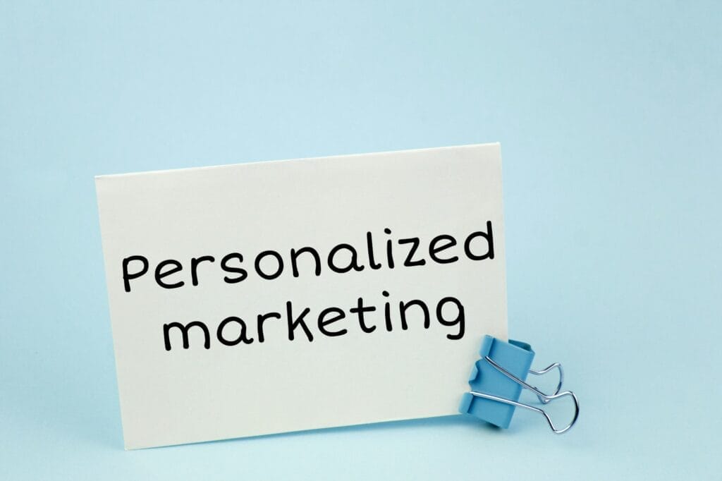 A white paper with the text "Personalized marketing" printed in black font is held up by a light blue binder clip. The background is a smooth and solid pale blue color. The image conveys the concept of personalized marketing in a simple and clear manner. © WEB6 Cloud